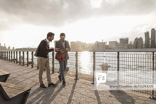 USA  New York City  zwei junge Männer  die am East River spazieren gehen und das Handy anschauen