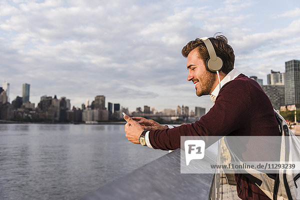 USA  New York City  lächelnder junger Mann mit Kopfhörer und Handy am East River