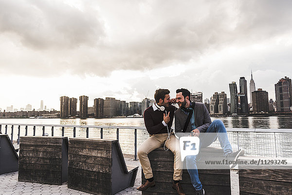 USA  New York City  zwei glückliche junge Männer mit Kopfhörer und Handy am East River