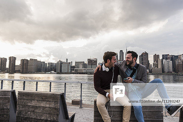 USA  New York City  zwei glückliche junge Männer mit Kopfhörer und Handy am East River