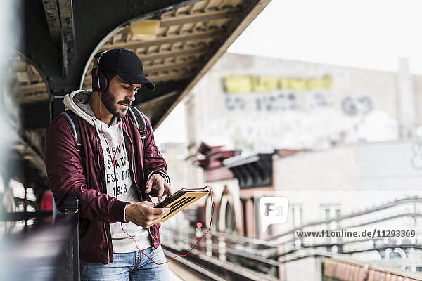 Junger Mann wartet auf U-Bahn  weraing Kopfhörer  mit digitalem Tablett