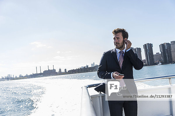 USA  New York City  Geschäftsmann auf der Fähre auf dem East River mit Handy und Kopfhörer