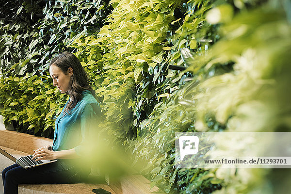 Junge Frau mit Laptop vor grüner Pflanzenwand