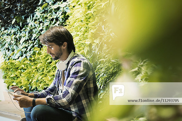 Junger Mann sitzt vor der grünen Pflanzenwand und benutzt eine digitale Tafel.