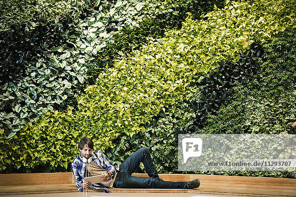 Junger Mann sitzt vor der grünen Pflanzenwand und benutzt eine digitale Tafel.
