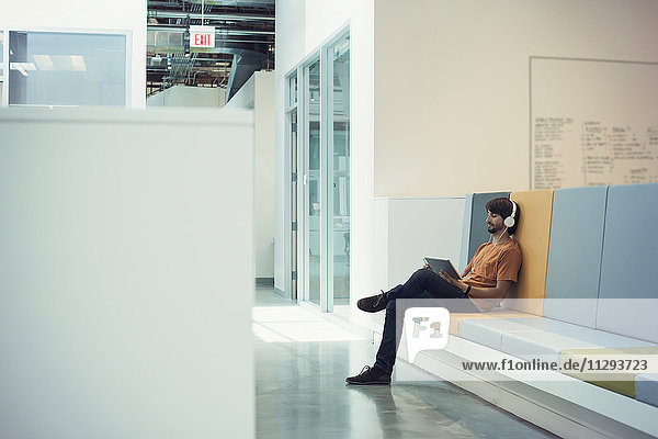 Junger Geschäftsmann mit Kopfhörer und digitalem Tablett  sitzend auf Bank mit