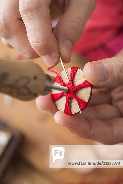 Knopfmacherin  Posamenten Knopf-Rohling aus Holz mit Haltewerkzeug wird mit rotem Garn von Händen umwickelt in Sternmuster und von hinten vernäht  dritter Schritt