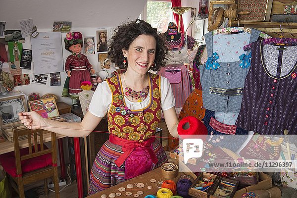 Button maker in colorful dirndl with Collier button trimmings in handicraft studio working with red yarn and utensils for Posamentenknöpfe  surrounded by traditional costumes  Ichenhausen  Bavaria  Germany  Europe