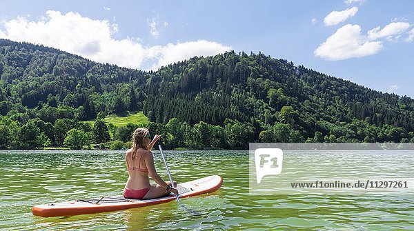 Junge Frau sitzt auf einem Stehpaddel Stand-Up-Paddel oder SUP Schliersee Oberbayern Bayern Deutschland Europa