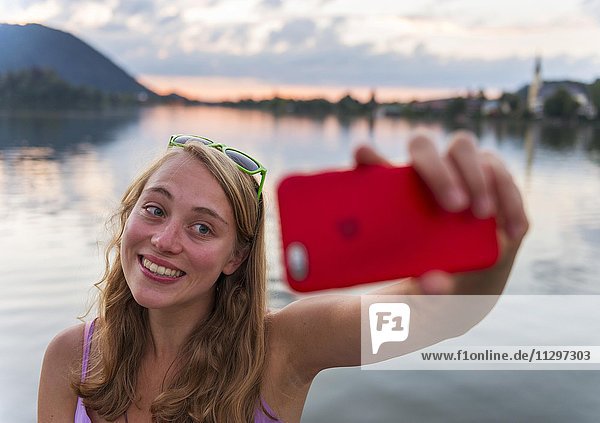 Junge Frau lächelt  fotografiert sich mit einem Handy  Selfie  Schliersee  Oberbayern  Bayern  Deutschland  Europa