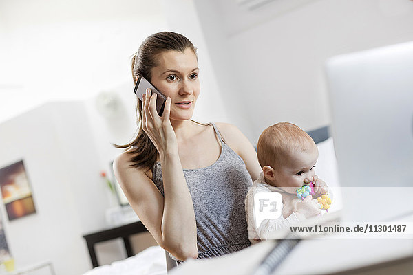 Mutter hält Baby-Tochter und arbeitet am Laptop und telefoniert mit dem Handy.