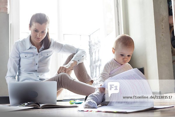 Curious baby daughter looking at paperwork next to mother working at laptop