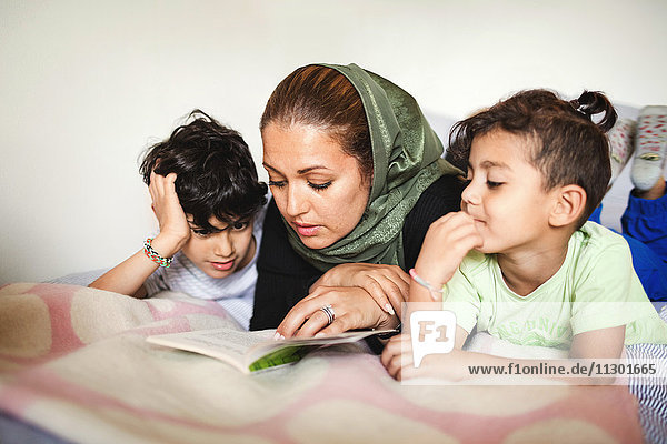 Mother reading book to boys while lying on bed at home