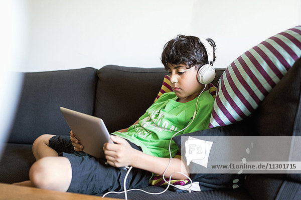 Junge  der Musik über ein digitales Tablett hört  während er zu Hause auf dem Sofa sitzt.