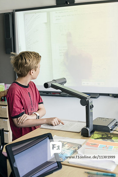 Boy reading document under camera while standing by desk in classroom
