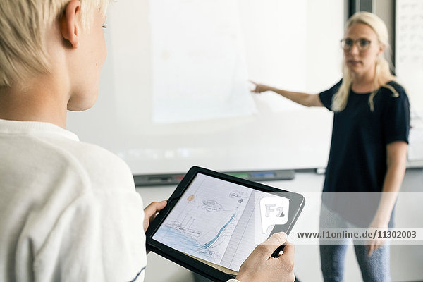 Beschnittenes Bild eines Jungen  der ein digitales Tablett hält  mit einem Lehrer  der im Klassenzimmer erklärt.