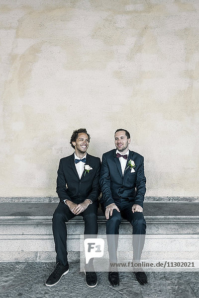 Happy newlywed gay couple sitting on bench against wall
