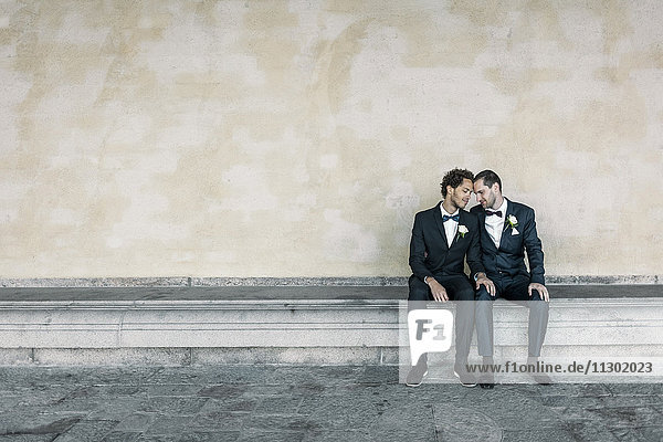 Neuvermähltes schwules Paar sitzt auf einer Bank an der Wand.