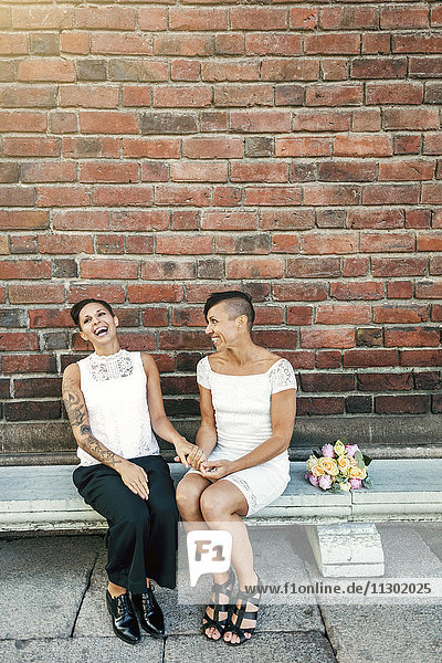 Fröhliches jungvermähltes lesbisches Paar auf der Bank an der Wand sitzend