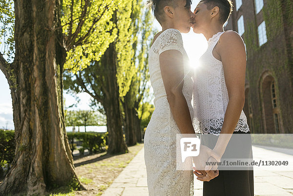 Seitenansicht eines frisch verheirateten lesbischen Paares  das sich küsst  während es sich auf dem Fußweg an den Händen hält.