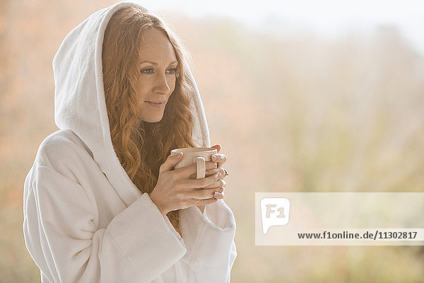 Gelassene Frau im Bademantel mit Kapuze trinkt Kaffee