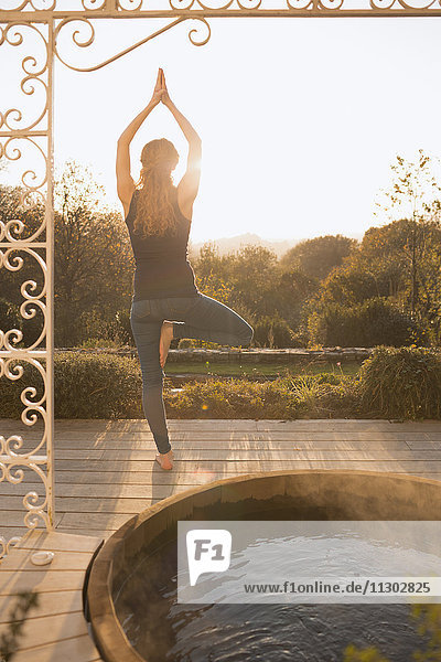 Frau übt Yoga-Baum-Pose auf Terrasse mit Whirlpool und Herbstbaumblick
