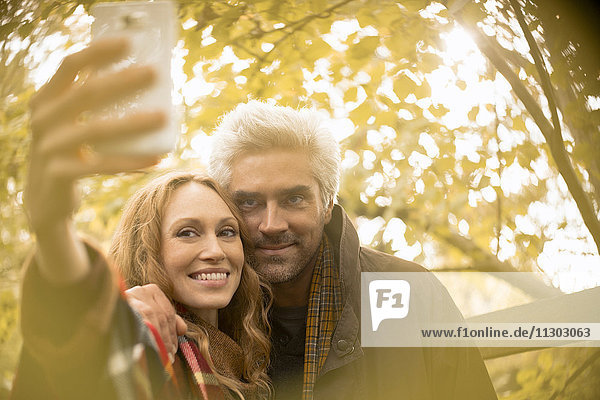 Smiling couple taking selfie with camera phone under autumn tree