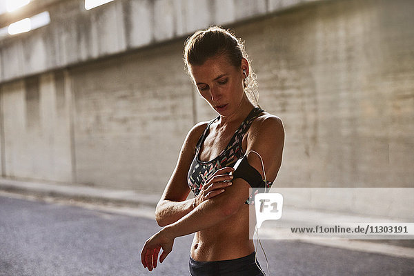 Fit female runner in sports bra using mp3 player arm band and headphones on urban street
