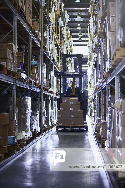 Worker driving forklift with cardboard boxes in aisle of distribution warehouse