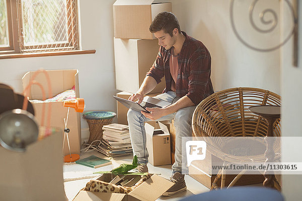 Junger Mann mit Laptop  umgeben von Umzugskartons in einer Wohnung