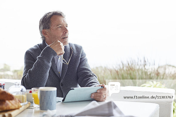 Nachdenklicher älterer Mann mit digitalem Tablet beim Frühstück auf der Terrasse