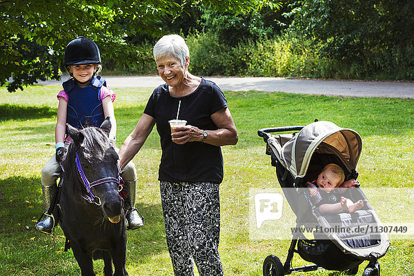 Woman  baby in pushchair and blond girl wearing riding hat sitting on a pony.