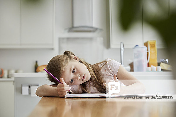 A girl sitting at a table in the family kitchen  holding a pencil  with her head in her hands.