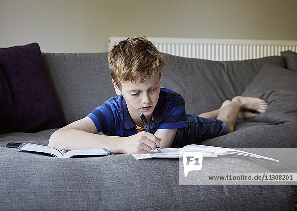 Ein Familienhaus. Ein Junge  der auf dem Sofa auf dem Bauch liegend seine Hausaufgaben macht und in ein Schulheft schreibt.