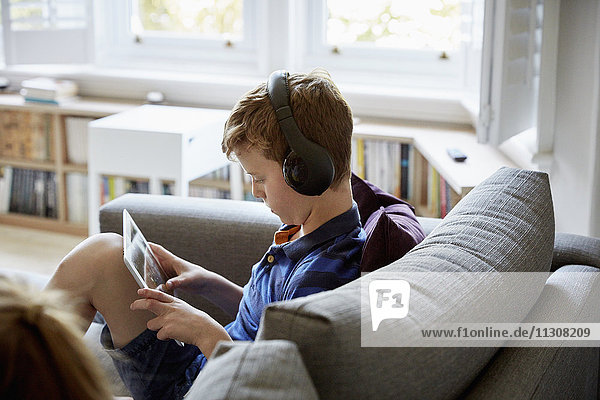 Ein Familienhaus. Ein Junge auf einem Sofa  der ein digitales Tablet benutzt und Kopfhörer trägt.