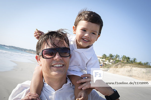 Porträt des Vaters  der seinen kleinen Sohn auf den Schultern am Strand trägt.