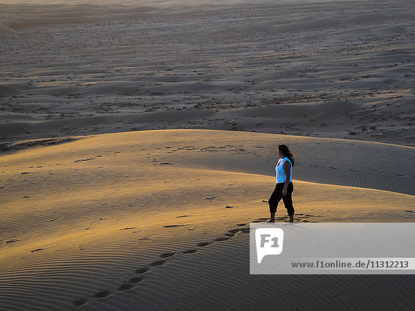 Oman  Al Raka  young woman standing on a desert dune in Rimal Al Wahiba desert watching sunset