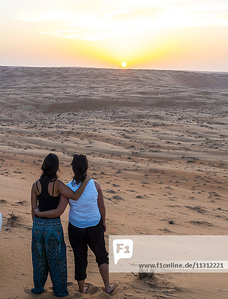 Oman  Al Raka  zwei junge Frauen  die Arm in Arm auf einer Wüstendüne stehen und den Sonnenuntergang beobachten.