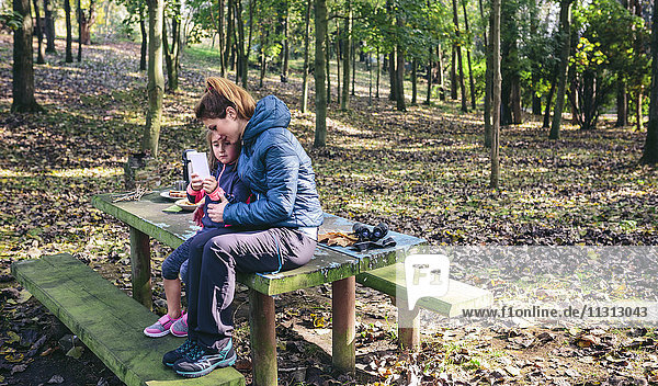 Mother and little daughter sitting at picnic place in the forest playing with smartphone