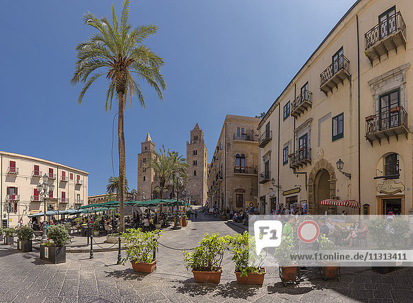 Square with outdoor cafes  sunshades and palm trees  Cefalu cathedral
