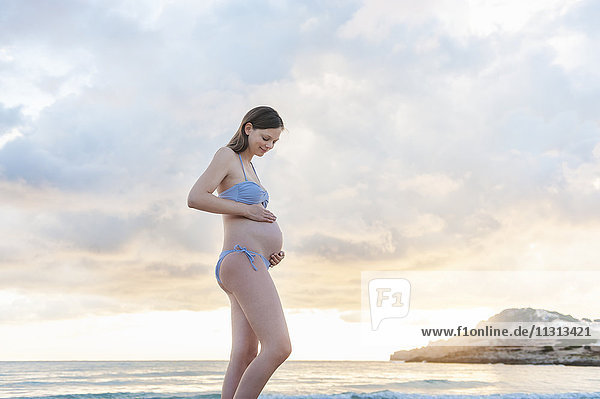 Schwangere Frau steht am Strand bei Sonnenaufgang