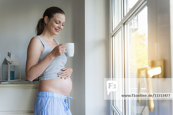 Smiling pregnant woman holding cup at the window