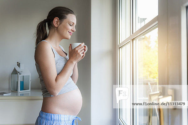 Lächelnde schwangere Frau hält Tasse am Fenster