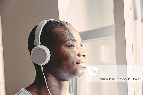 Porträt eines lächelnden jungen Mannes  der mit Kopfhörern Musik hört  während er durchs Fenster schaut.