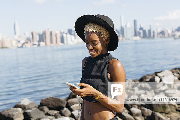 USA  New York City  Brooklyn  lächelnde junge Frau am East River mit Blick aufs Handy