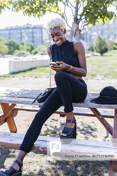Lächelnde junge Frau sitzt auf einem Holztisch und schaut aufs Handy.