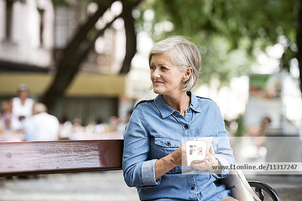 Seniorin mit Smartphone sitzt auf der Bank in der Stadt