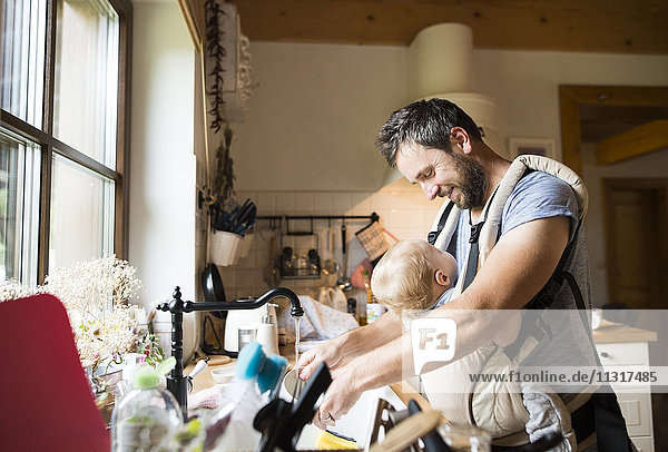 Happy father with baby in baby carrier doing the dishes