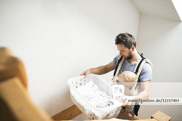 Father with baby in baby carrier carrying laundry basket walking upstairs
