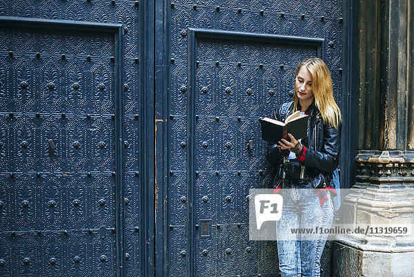 Spain  Barcelona  young woman standing in front of blue entrance gate reading a book